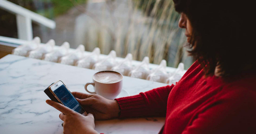 A woman sits at a table with her coffee and looks at her cell phone.