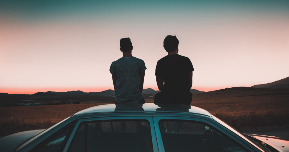 Two men sit on the roof of a car looking at a sunset