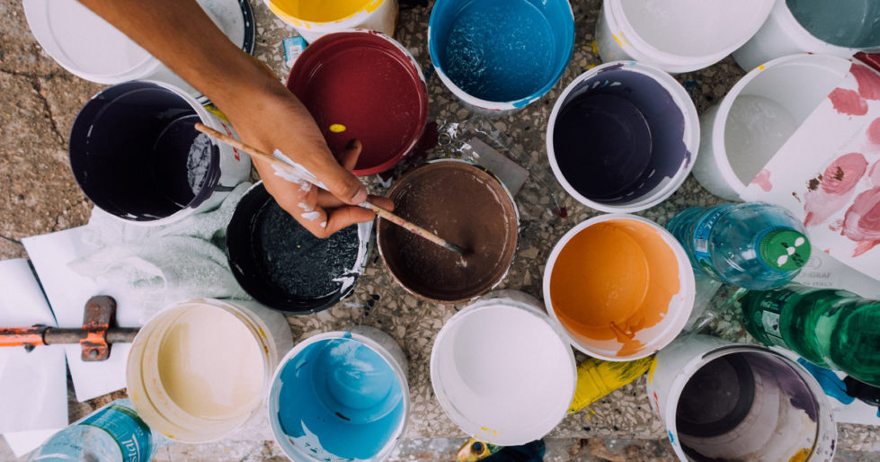A hand holding a paintbrush dips it into a pail of brown paint, with many other colours and pails of paint surrounding it.