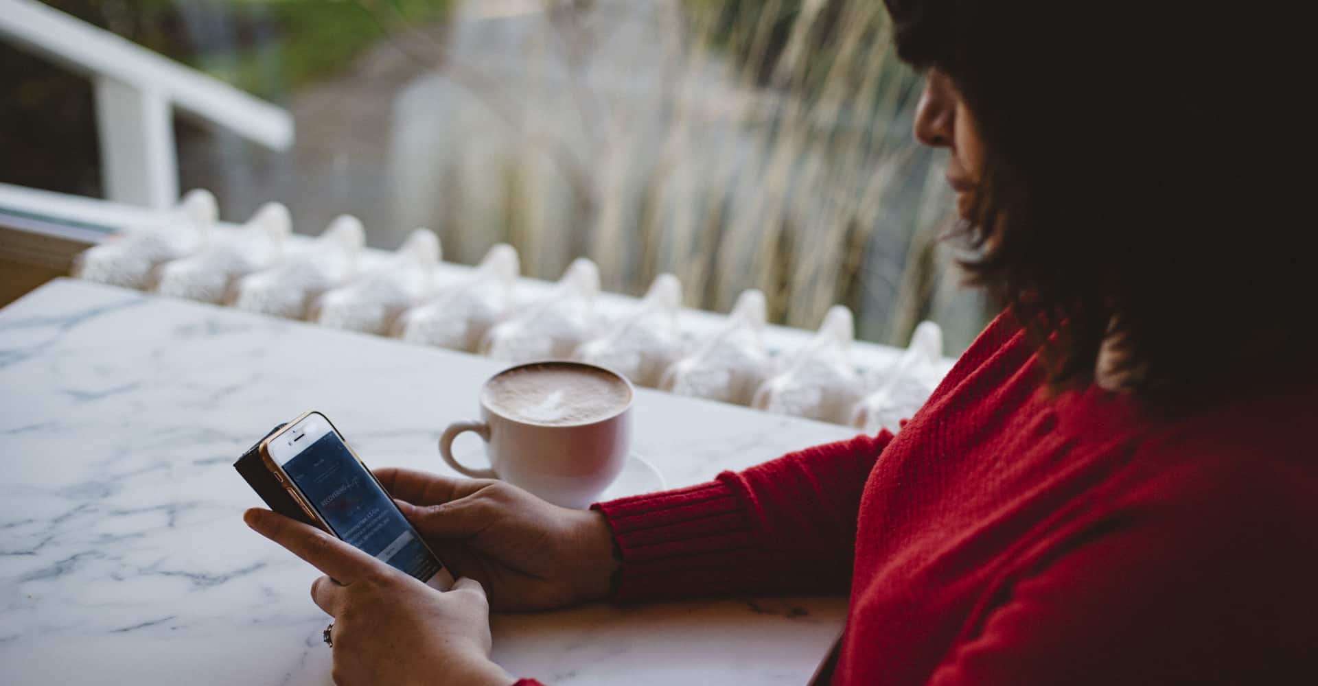 A woman sits at a table with her coffee and looks at her cell phone.