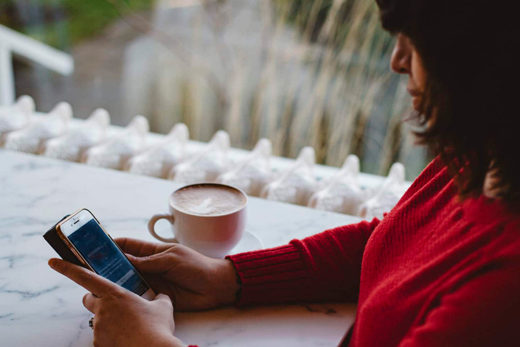 A woman sits at a table with her coffee and looks at her cell phone.