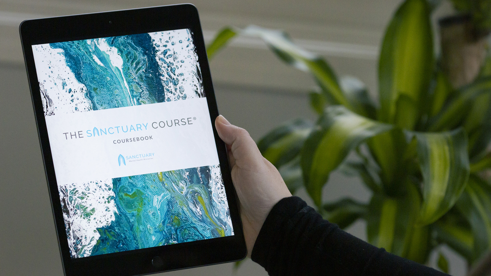 A hand holds a tablet showing The Sanctuary Course Coursebook cover